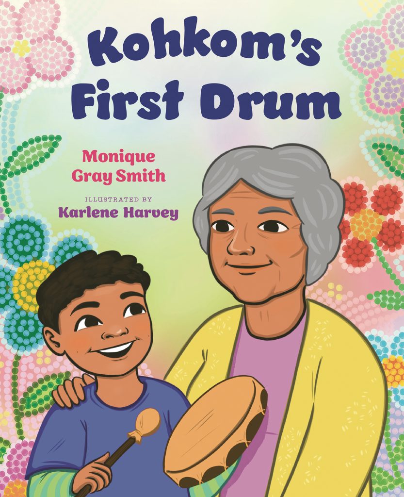 Kohkom's First Drum by Monique Gray Smith. Illustrated by Karlene Harvey. A boy holding a hand drum smiles up at his grandma, who has her arm around his shoulder. Behind them is a colourful beadwork design.